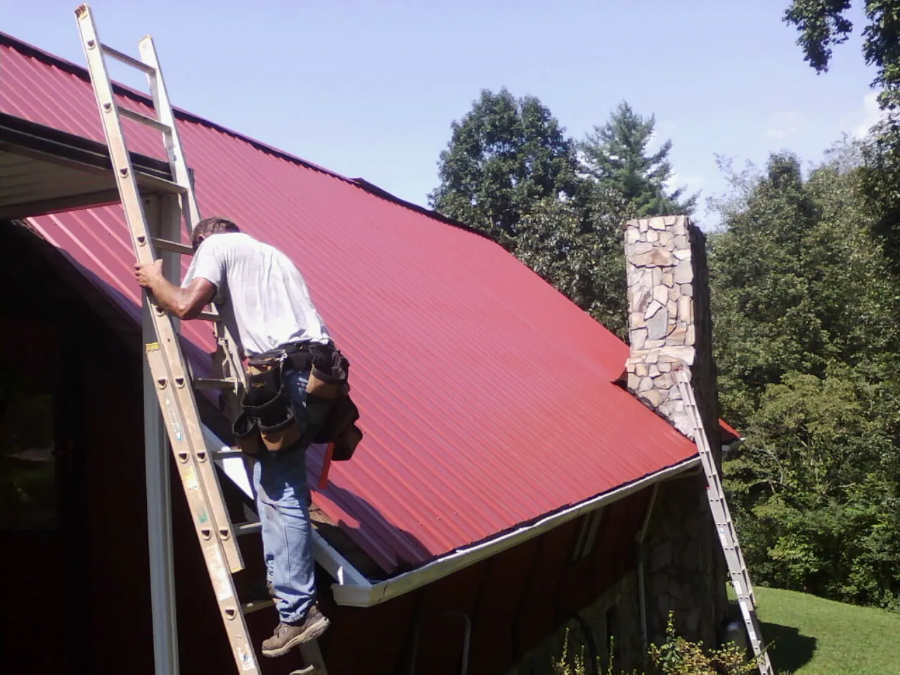 Roofer on a ladder beside a red metal roof and stone chimney — Soffit & Fascia Repair by Roofing Brothers in Knox